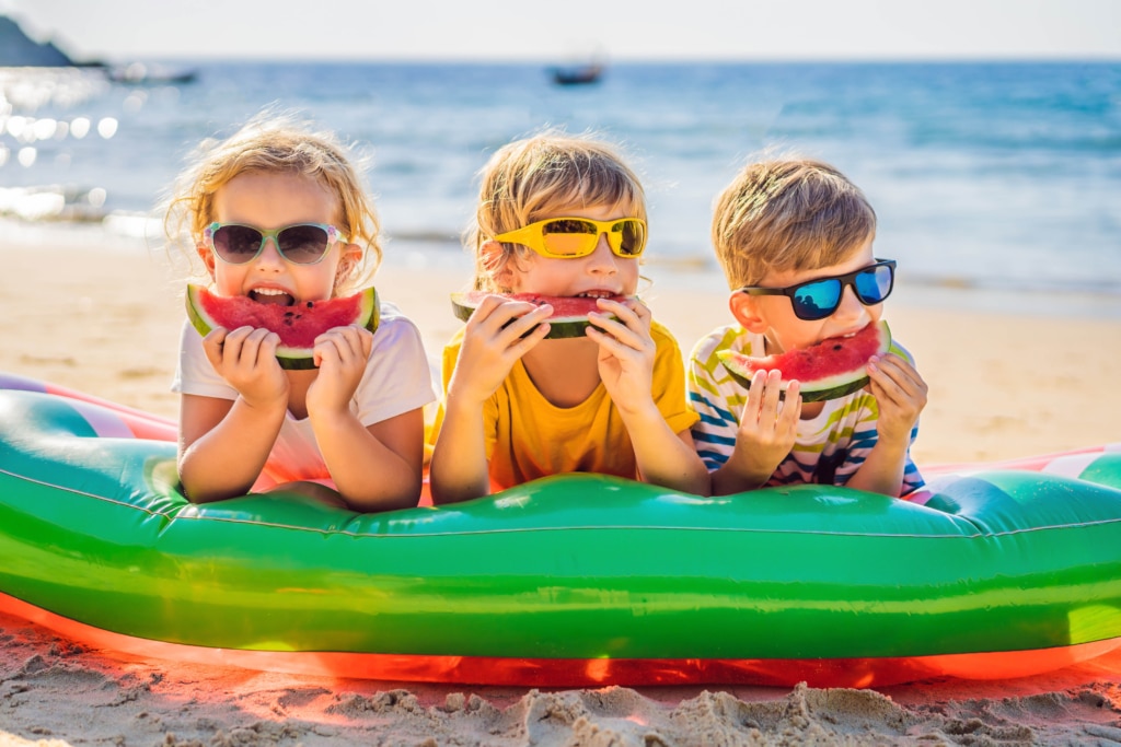 Tre små barn som spiser vannmelon i vannkanten en fin og trygg sommerdag